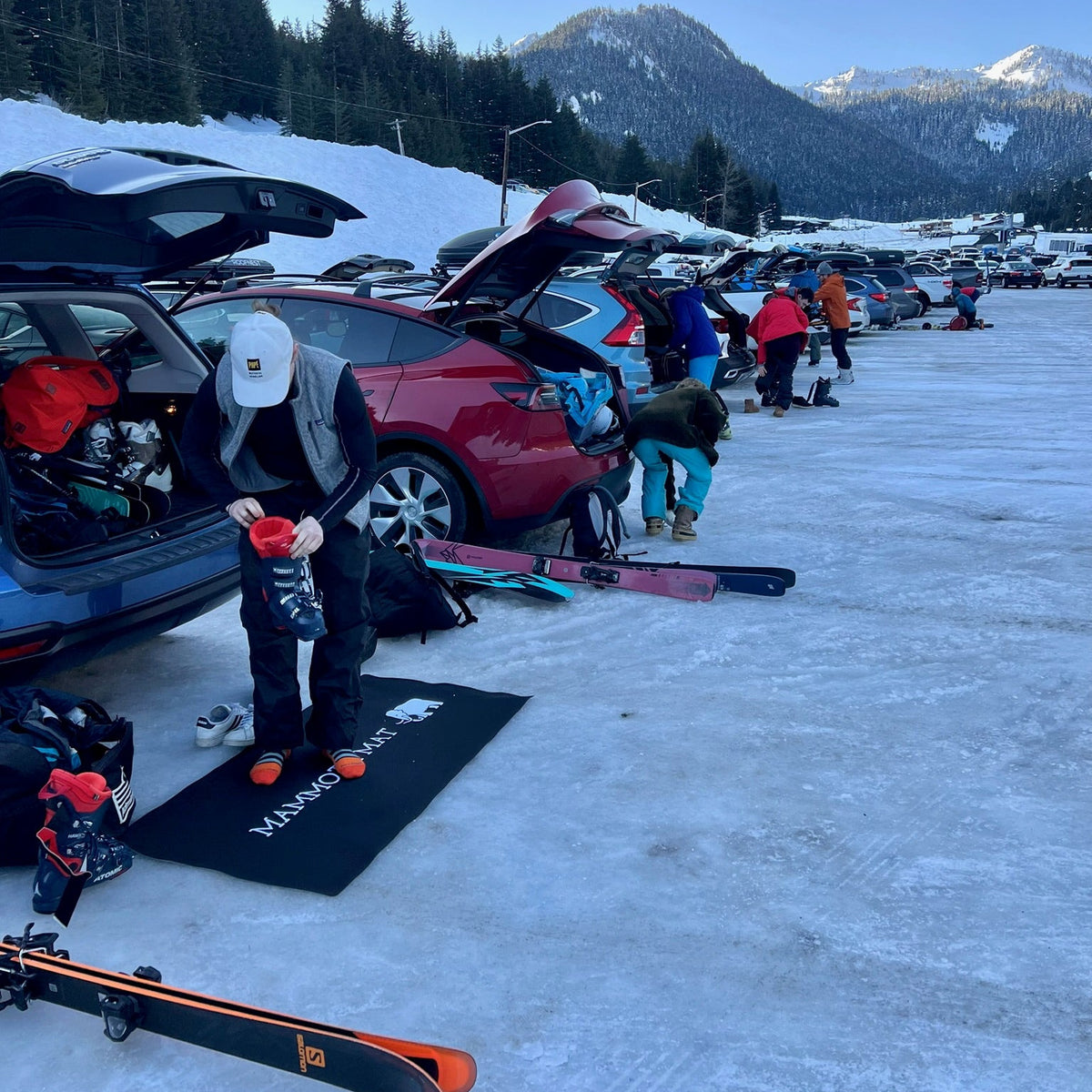 People preparing for skiing with cars and skis in a snowy parking lot with mountains in the background. Waterproof ski mat makes it easier for this person to change into their boots.